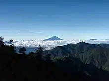 Photo couleur de la silhouette du sommet conique d'une montagne, émergeant d'une mer de nuages blancs au loin (centre de la photo). Un massif montagneux boisé, au premier plan et un ciel bleu en arrière-plan.