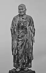  Muchaku (Asanga). Vue de face d'une statue réaliste gardant les mains en face de son corps avec une paume tournée et l'autre de côté, comme pour conserver quelque chose. Photographie noir et blanc.