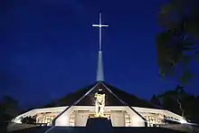 Photo nocturne d'une église en forme de cône large et aplati, surmontée d'une haute croix, derrière une statue représentant André Kaggwa mutilé.