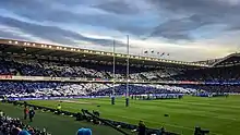 Photographie en couleurs panoramique des gradins d'un stade, avec les spectateurs brandissant des cartons blancs ou bleus pour que l'ensemble constitue le drapeau écossais.