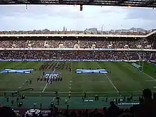 Photographie en couleurs. Au centre d'un terrain de rugby, un orchestre musical en représentation, entouré par deux drapeaux écossais géants étendus au sol. En arrière-plan, des spectateurs sont en place dans une tribune.
