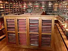 Grande salle aux murs couverts d'étagères de livres, parcourues par une galerie. Au centre, un grand meuble en bois contenant de grands registres