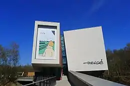 Vue d'ensemble du musée depuis la passerelle d'accès. Une case d'un album est reproduite sur une façade à gauche, et la signature d'Hergé à l'opposé.