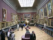 Grande salle à éclairage zénithale couverte de tableaux avec une grande porte centrale et une dizaine de visiteurs.