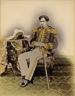 Photo noir et blanc d'un homme assis sur un fauteuil, en tenue impériale officielle, un sabre à la hanche, un bicorne posé sur une table à gauche.