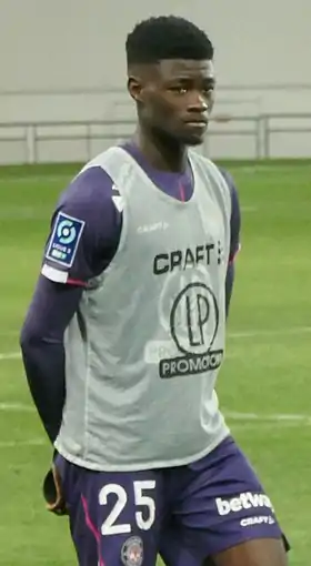Plan trois-quarts de Steve Mvoué, short et maillot violet, chasuble blanche, bras derrière le dos