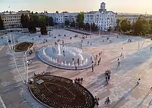 Photographie en couleurs. Une fontaine ovale se dresse au centre de la place avec, en contre-haut et en contre-bas à gauche, deux autres ovales végétalisés de surface similaire. La place est parcourue par de nombreux flâneurs. Présence de nombreux éclairages publics éteints. Des bâtiments bordent la place. Le ciel est dégagé et partiellement orangé.