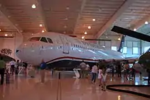Photo prise à l'intérieur d'un hangar montrant l'avion endommagé exposé dans un musée. Des visiteurs sont présents tout autour et admire l'avion.