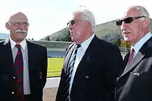 Trois hommes en costume cravate discutent près d'un stade de rugby à XV.