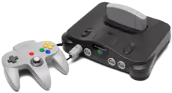On the left, a three-pronged, handheld controller with a central analog stick and multiple buttons. On the right, a black electronics unit that accepts a light gray cartridge on the top and controllers (via ports) on its front.
