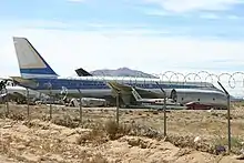 Photo couleur d'un avion blanc avec des bandes bleues, prise devant un grillage avec des barbelés. Au milieu du désert, il repose avec de plus petits avions autour.