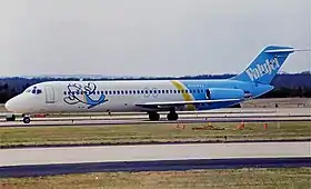Photo du côté gauche d'un avion de ligne aux couleurs blanches à l'avant et bleues à l'arrière, roulant sur un aéroport. Les deux moteurs sont situés à l'arrière et un dessin enfantin d'un avion, souriant et levant les ailes en guise de bras, est présent sur l'avant du fuselage.