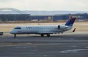 Photo de trois quart face d'un avion de ligne, aux couleurs blanches avec des inscriptions bleues et rouges, à la porte d'embarquement d'un aéroport avec la passerelle passagers contre la porte opposée de l'avion.