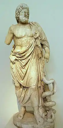 Statue du dieu grec Asclépios, debout. Barbu, il est habillé avec une toge qui laisse son torse nu. À sa gauche, un serpent est enroulé autour d'un bout de bois. La statue est fêlée à plusieurs endroits, il manque des doigts à sa main gauche et son avant-bras droit. Sa main droite est posée contre sa hanche.