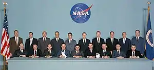 Photographie en couleur du groupe d'astronautes 5 : les hommes en costume sont alignés sur deux rangées avec au premier plan des maquettes de vaisseaux spatiaux.