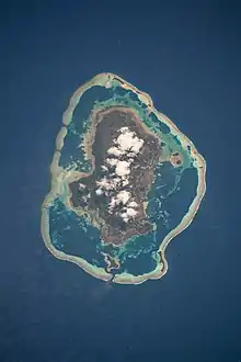 île brune et blanche au centre d'un lagon formé par une barrière de corail circulaire, le tout entouré de mer bleue