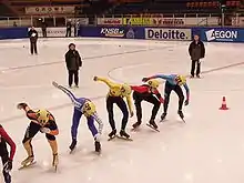 Cinq patineurs alignés sur la ligne de départ, avec deux juges derrière et sur le côté.