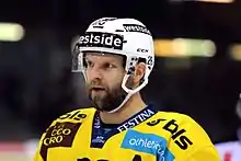 Photographie couleur d’un hockeyeur avec un maillot jaune et un casque blanc, barbu, dont on ne voit que le buste