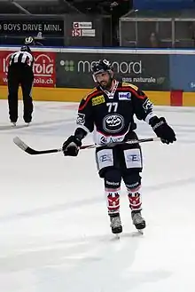 Photographie couleur d’un hockeyeur, en pied, de face, tenant sa canne horizontalement dans ses mains, à hauteur de hanche, la tête et le regard légèrement penché sur sa droite.