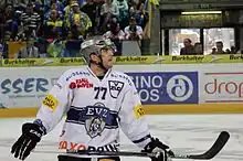 Photographie couleur d’un hockeyeur de face, vêtu de blanc, la tête tournée sur sa gauche, les yeux en l’air