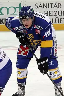 Photographie couleur d’un hockeyeur en pied, penché en avant, sa canne posée sur la glace sur sa gauche