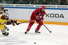 Au centre de l’image, un hockeyeur tout de rouge vêtu, de pied ; à l’extrême gauche, un autre, vêtu à dominante jaune, très penché en avant, voulant gêné le premier avec sa canne.