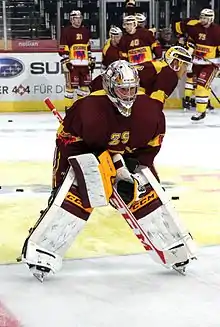 Photographie couleur d’un gardien de but de hockey sur glace, en pied, jambes écartées, légèrement penché en avant, au milieu de la patinoire