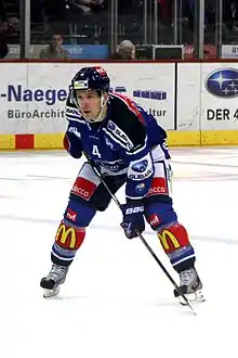 Image couleur d’un hockeyeur penché en avant, les jambes écartés, vêtu principalement de bleu.