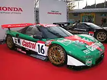 Photographie d'une voiture de GT vert foncé, rouge et blanche, vue de trois-quarts gauche, exposée dans un stand d'exposition, sur un sol rouge.