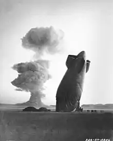 The tail, or “aft”, section of a U.S. Navy Goodyear ZSG-3 (en) Blimp is shown with the Stokes cloud in background. The blimp was over five miles from ground zero when it was collapsed by the shock wave. The airship was unmanned and was used in military effects experiments on blast and heat. Navy personnel on the ground in the vicinity of the experimental area were unhurt. On the ground to the left are the re mai ns of the blimp's forward section.