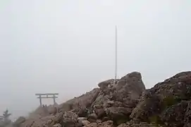 Photo couleur d'une lame de sabre enchâssée dans une roche au sommet d'une montagne, avec un ciel brumeux en arrière-plan.