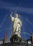 Statue blanche, sur fond de ciel bleu, encadrée par des jets d'eau