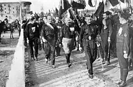 Photographie en noir et blanc de Benito Mussolini, au deuxième plan à gauche de l'image, lors de la marche sur Rome