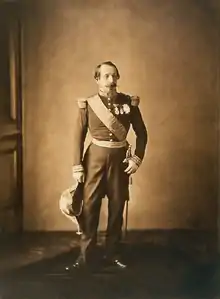 Photographie en sépia de Napoléon trois, de plain pied. Il porte un uniforme militaire et tient un bicorne de sa main droite. Son visage arbore un air éteint, comme sur de nombreuses photographies.