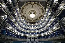 Vue depuis la scène des balcons d'un théâtre à l'italienne, puissamment éclairés. La tonalité de l'ensemble est bleutée.