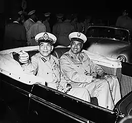 Photographie de deux hommes souriants en uniformes militaires assis à l'arrière d'une voiture décapotable.