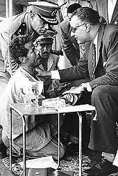 Un homme à genoux regarde un homme assis qui lui tient la main. Plusieurs hommes en uniforme militaires se tiennent à l'arrière-plan.