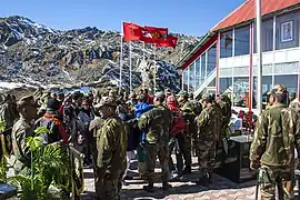 Scène de cérémonie avec soldats indiens en treillis et civils à proximité d'un bâtiment vitré au toit rouge.