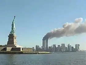 La Statue de la Liberté au premier plan avec les deux tours du World Trade Center en feu et dégageant une épaisse fumée noire.
