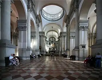 Image de l'intérieur d'une nef d'église avec des colonnades et arches de chaque côté, un pavement à damier, une coupole en hauteur et au fond un autel.