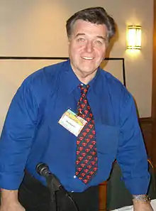 Photo en buste d'un homme souriant, âgé d'une soixantaine d'années, portant une chemise bleue et une cravate, devant lui un micro