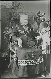 Une dame âgée, assise dans un fauteuil, entourée de fleurs et de feuilles.