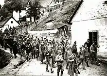 Des prisonniers, encadrés par des hommes en armes, marchant à travers un village.