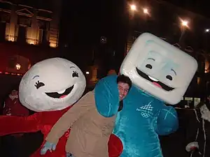 Photographie de deux mascottes, l'une en rouge à gauche, l'autre en bleu à droite, encadrant un homme jeune.