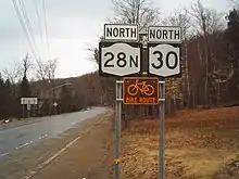 Signage for Route 28N and Route 30 heading northbound through a wooded community with a brown and white bicycle route sign