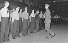 Photo en noir et blanc d'hommes en civil se tenant en rang cc un homme en uniforme militaire portant une casquette et un bâton à la main se tenant devant eux à l'intérieur d'un bâtiment