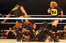 Sur un ring, trois membres du Nexus — Justin Gabriel (de dos), Wade Barrett (masqué) et Heath Slater — attaquent le catcheur Goldust, à terre.