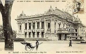 Photo ancienne représentant un grand théâtre d'architecture occidentale. On aperçoit devant le bâtiment des passants coiffés de chapeaux chinois.