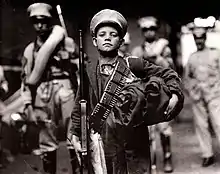 Photo en noir et blanc d'un enfant coiffé d'une casquette de soldat, un fusil dans les mains et une cartouchière en bandoulière.