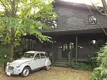 Photographie d'une maison en bois entourée de végétation devant laquelle est garée une voiture blanche plutôt ancienne.